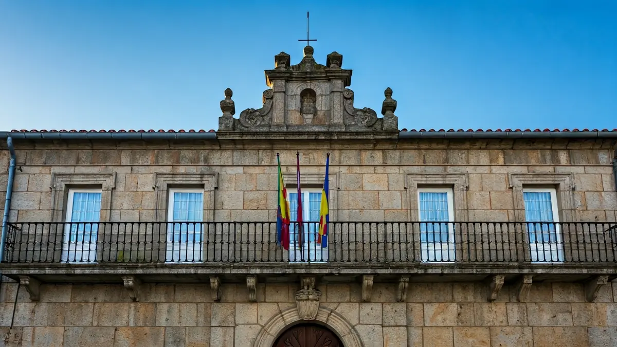 Imaxe da fachada dun concello galego, con balcón e varandas de ferro, baixo a luz do sol da tarde.