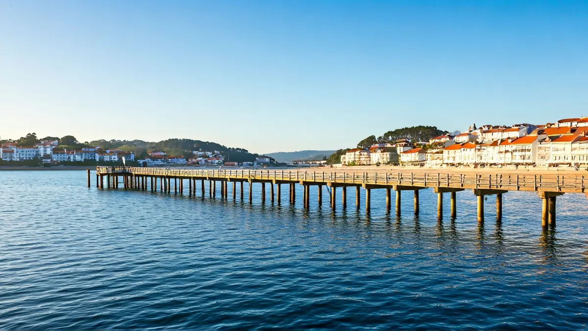 Imaxe dun peirao de madeira nunha praia galega, con casas tradicionais e augas tranquilas.