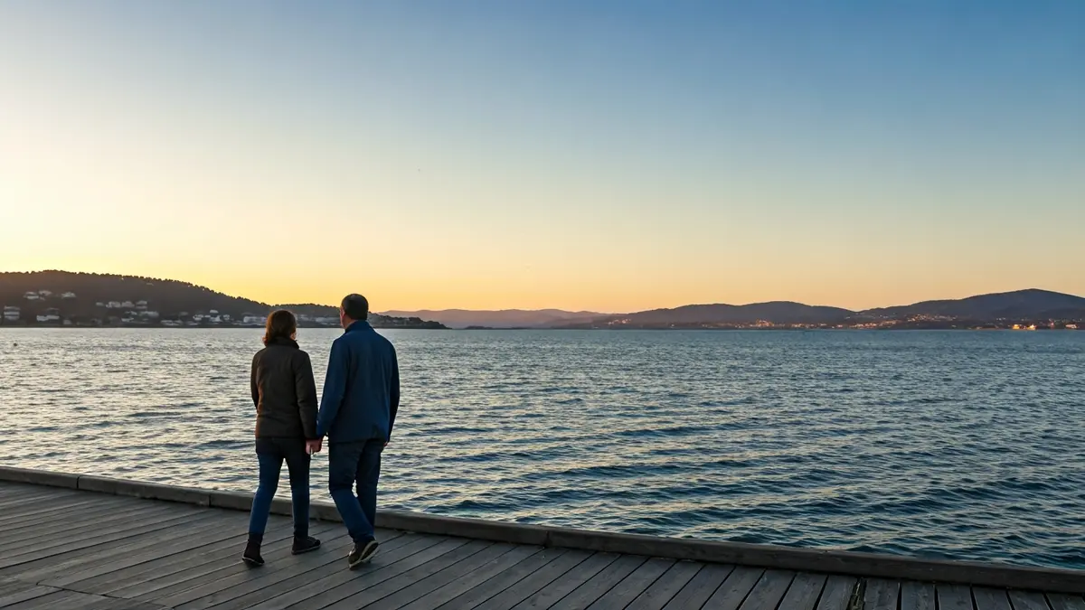 Imaxe dunha parella camiñando por un peirao ao atardecer, co mar en calma e a costa galega ao fondo.