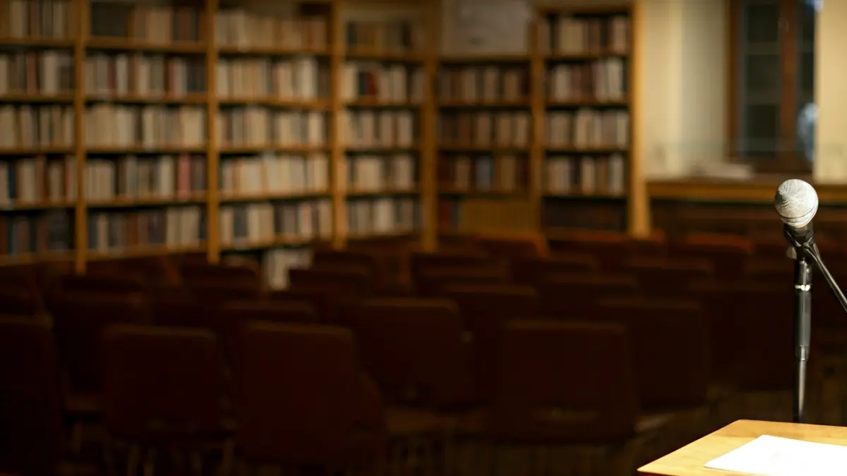 Imaxe xenérica dunha sala de conferencias ou biblioteca, con cadeiras e un atril con micrófono.