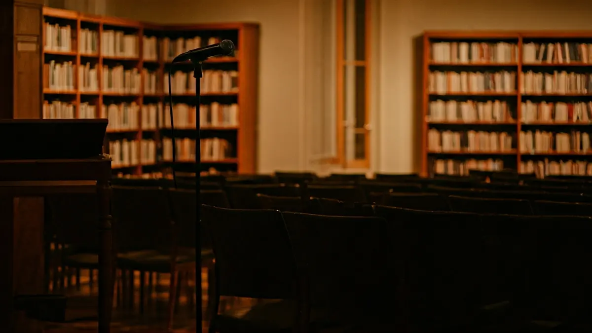 Imaxe xenérica dunha biblioteca con estantes de madeira e un micrófono nun podio, con cadeiras baleiras