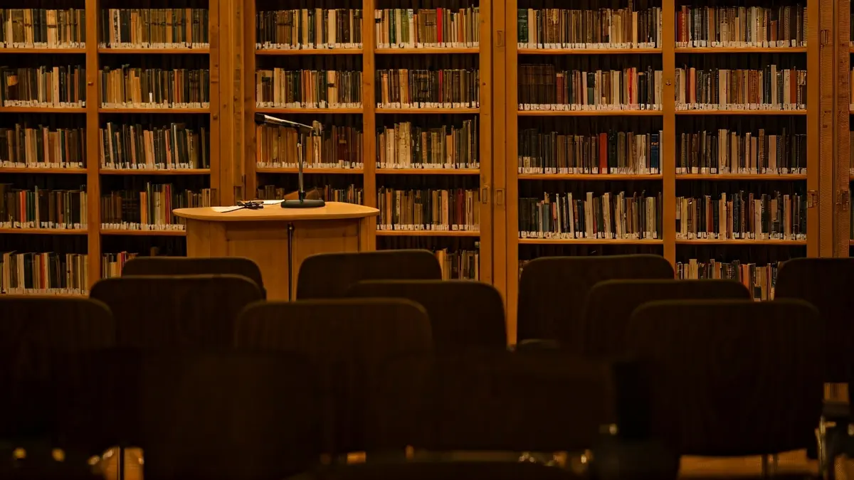 Imagen genérica de una sala de presentaciones o biblioteca, con estantes de madera y un micrófono en un atril.