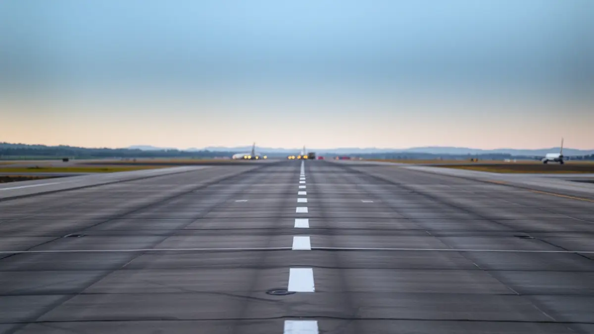 Imaxe dunha pista de aeroporto baleira ao amencer, con vehículos de mantemento ao lonxe.