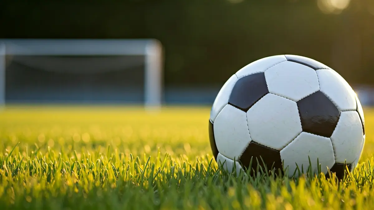 Imaxe xenérica dun balón de fútbol sobre a herba dun campo, coa portería difuminada ao fondo.
