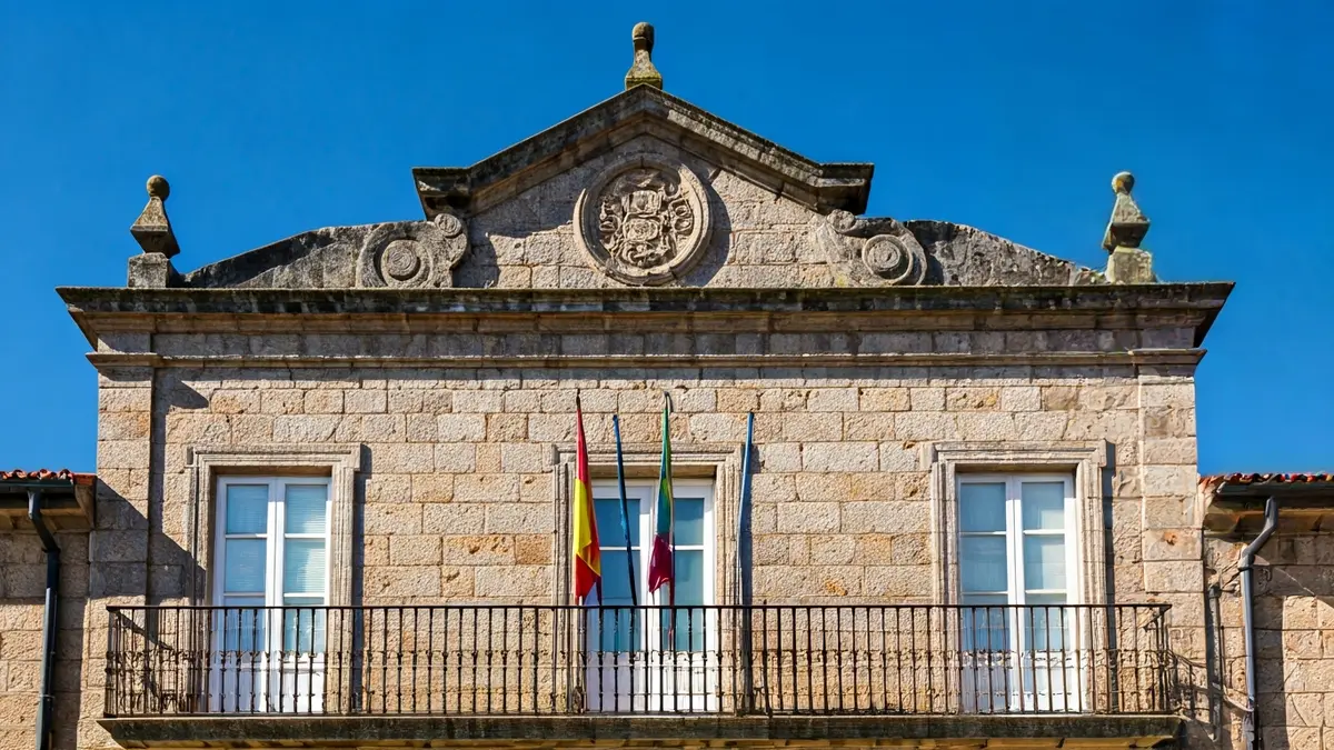 Imaxe da fachada dun concello galego, con balcón e varandas de ferro, baixo a luz do sol da tarde.