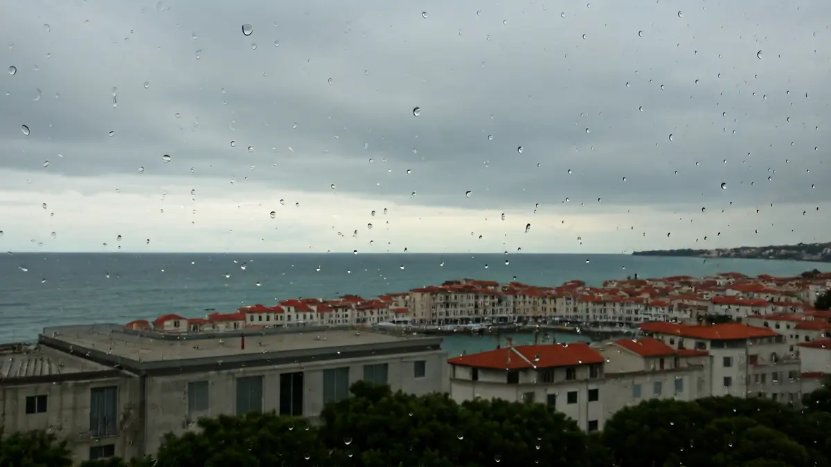 Imaxe xenérica dun ceo anubrado sobre unha vila costeira, con pingas de choiva nun cristal.