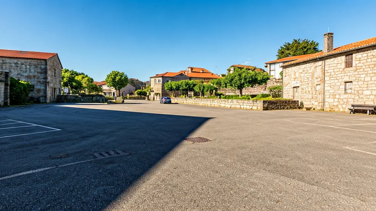Imaxe dun aparcadoiro baleiro nunha vila galega, rodeado de edificios de pedra.