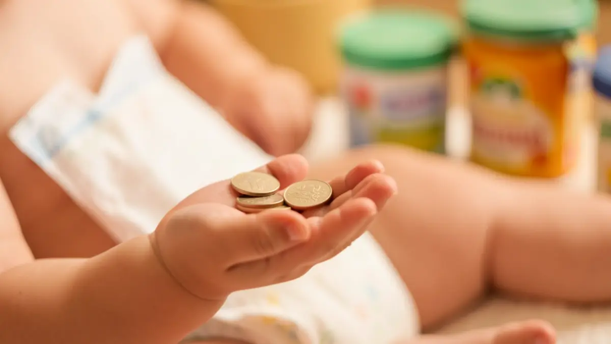 Primeira imaxe dun bebé sostendo moedas, con produtos de hixiene e alimentación infantil desenfocados ao fondo.