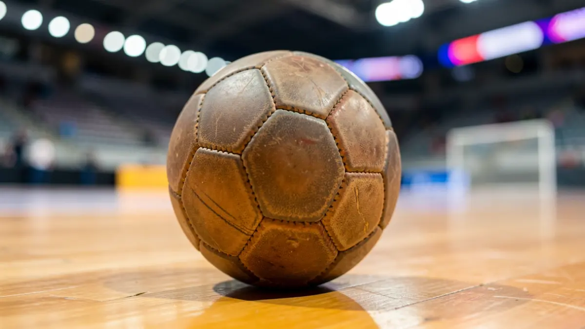 Imagen genérica de un balón de balonmano sobre una cancha de madera pulida, con luces de estadio desenfocadas al fondo.