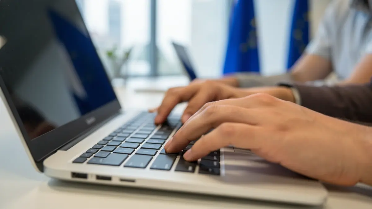 Imaxe xenérica de mans diversas escribindo nun teclado de portátil nunha oficina moderna con motivos da Unión Europea.