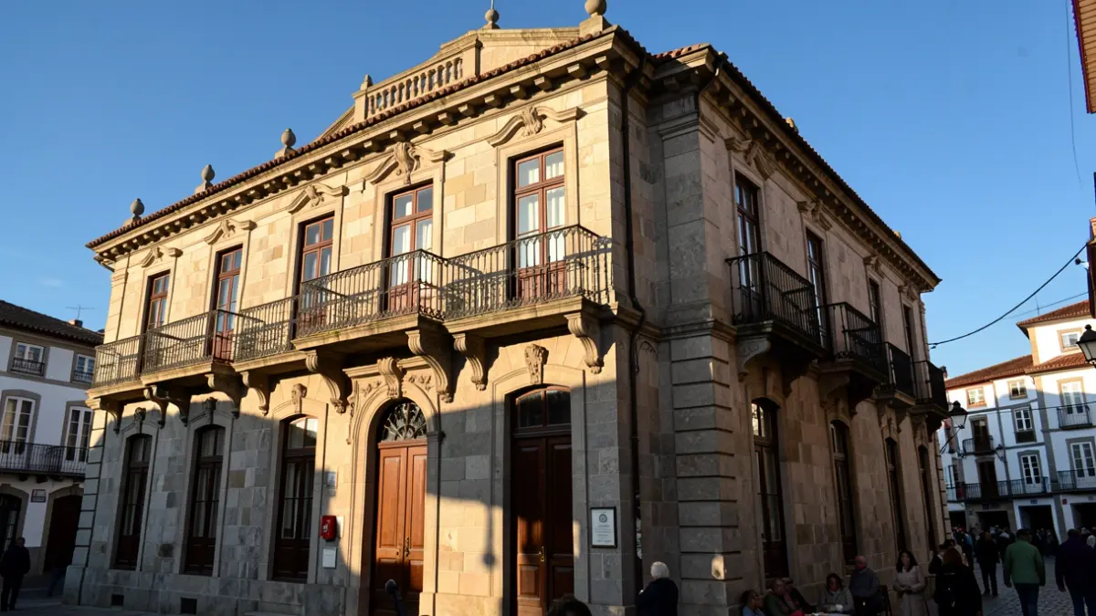 Imaxe xenérica dun concello galego de granito con balcóns ornamentados e reixas de ferro, á luz da tarde.